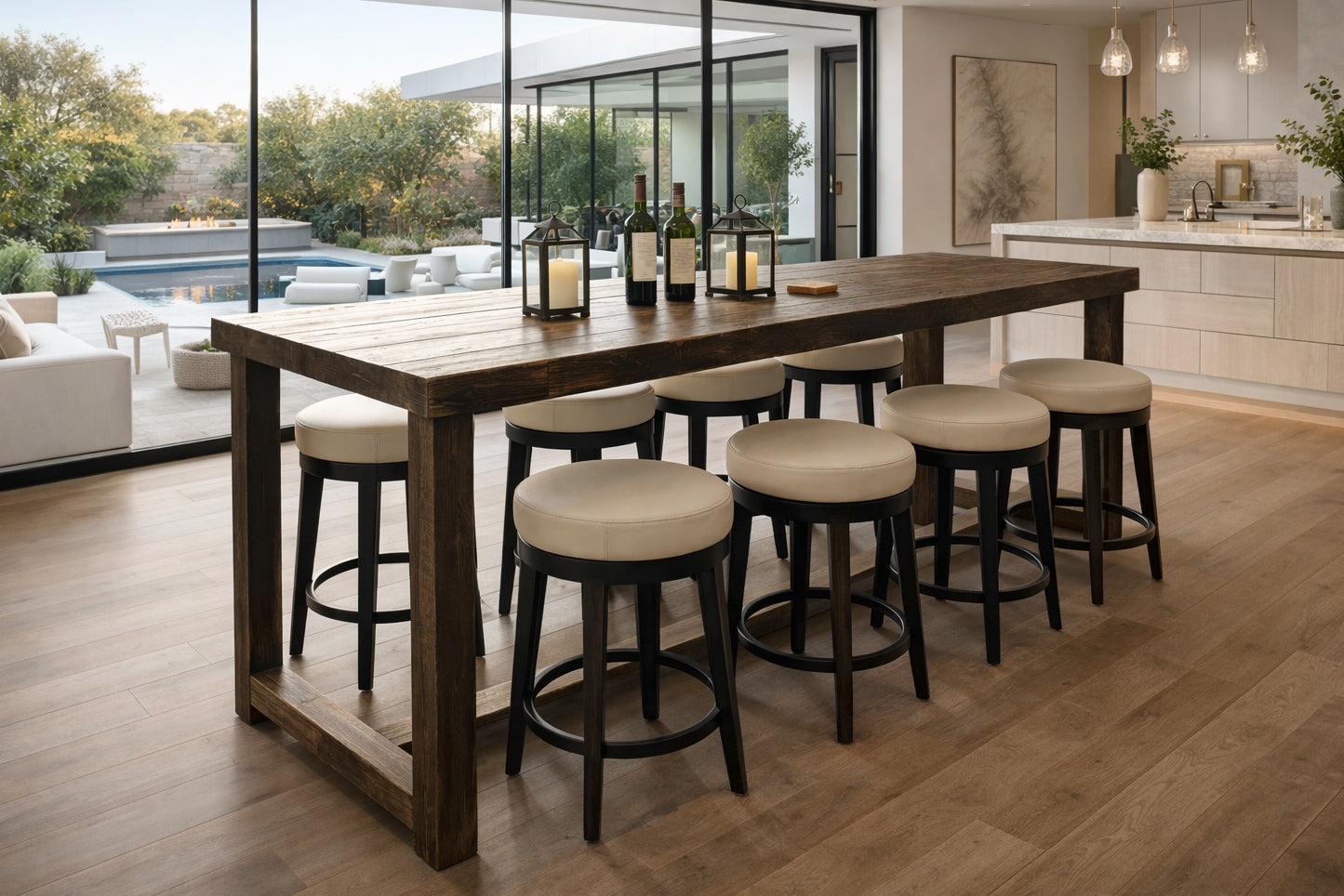 This inviting kitchen showcases a handcrafted reclaimed wood kitchen island, bar-height table style, paired with six cushioned stools. Elegant lanterns and wine bottles create warmth, while large windows frame a serene patio and pool view.
