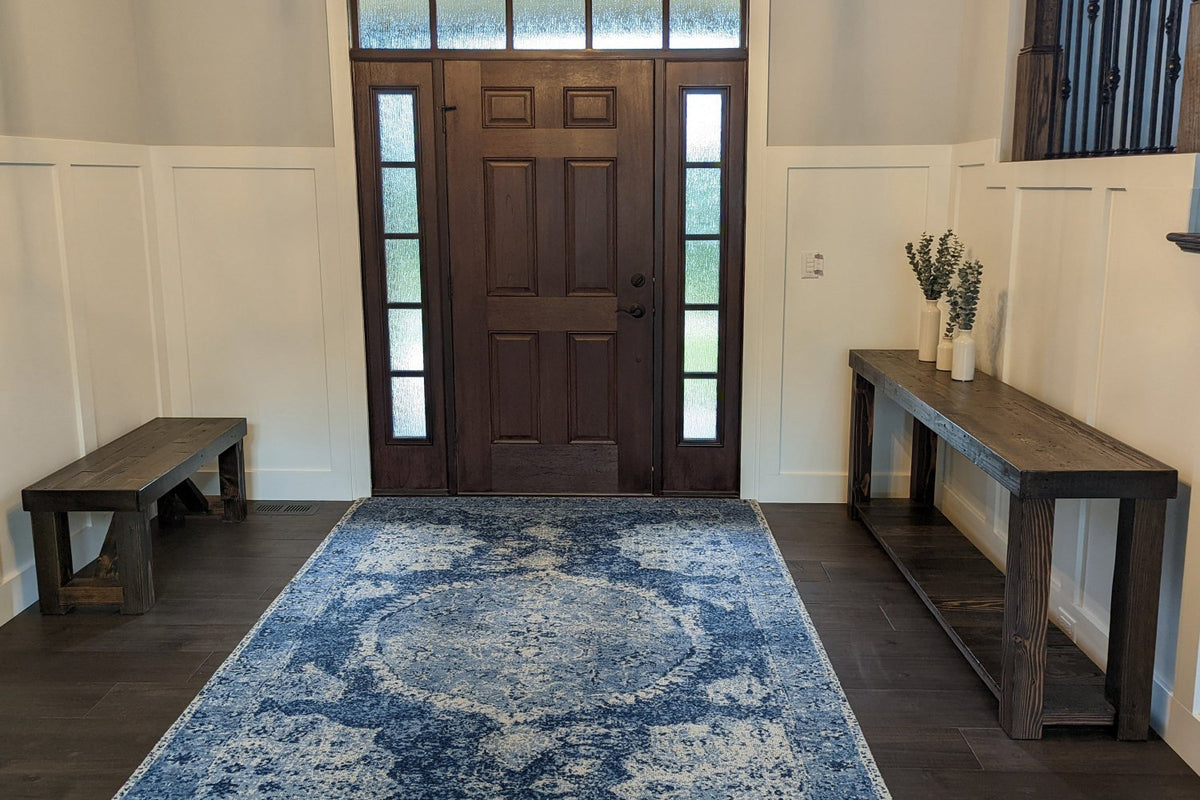 A handcrafted reclaimed wood console table, expertly made in Arizona, stands to the right of a wooden front door with frosted glass panels. This narrow table features vases and greenery, complementing sustainable style and quality.