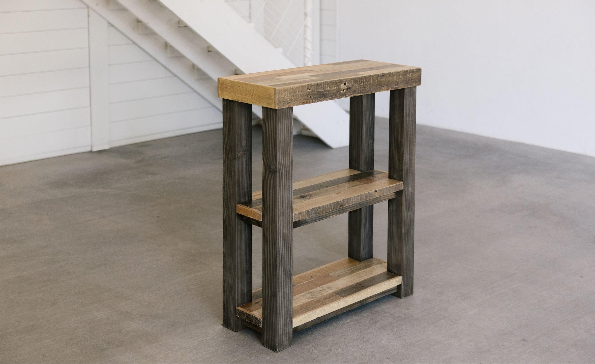 Handcrafted reclaimed wood console table with three shelves and natural finish on concrete floor by white staircase; ideal for modern café, minimalist space, or historic home. Sustainable furniture made in Arizona.