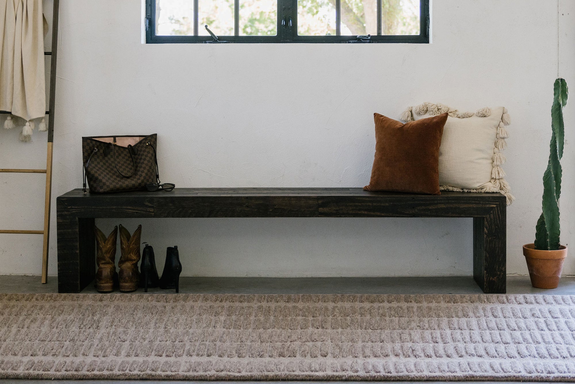 Handcrafted in Arizona, this reclaimed wood bench showcases enduring quality and sustainability. Two plush pillows, a handbag, and shoes add livable warmth; a potted cactus completes the minimalist entryway furniture.