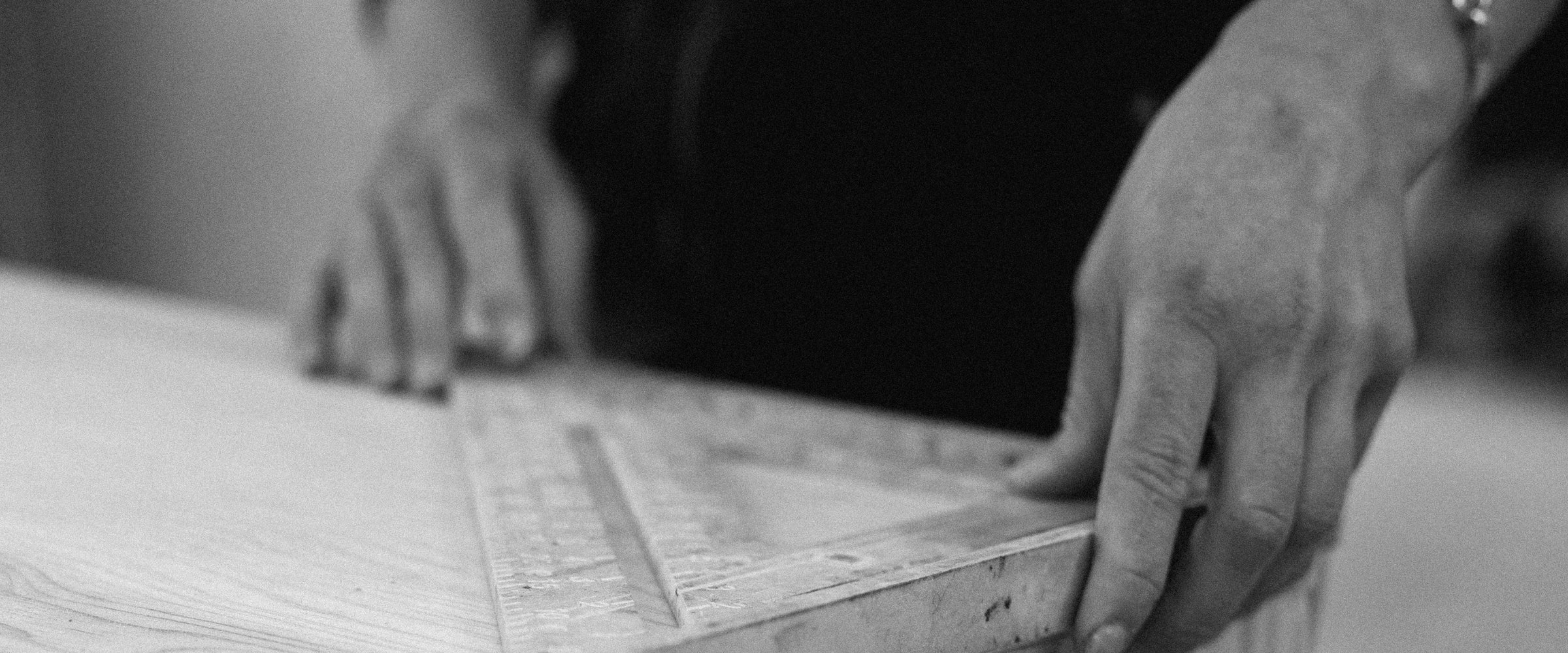 A close-up, black-and-white photo showcases skilled hands using a metal carpenter’s square to mark reclaimed wood on a workbench—highlighting the attention to detail and quality craftsmanship behind our handcrafted Arizona dining tables.