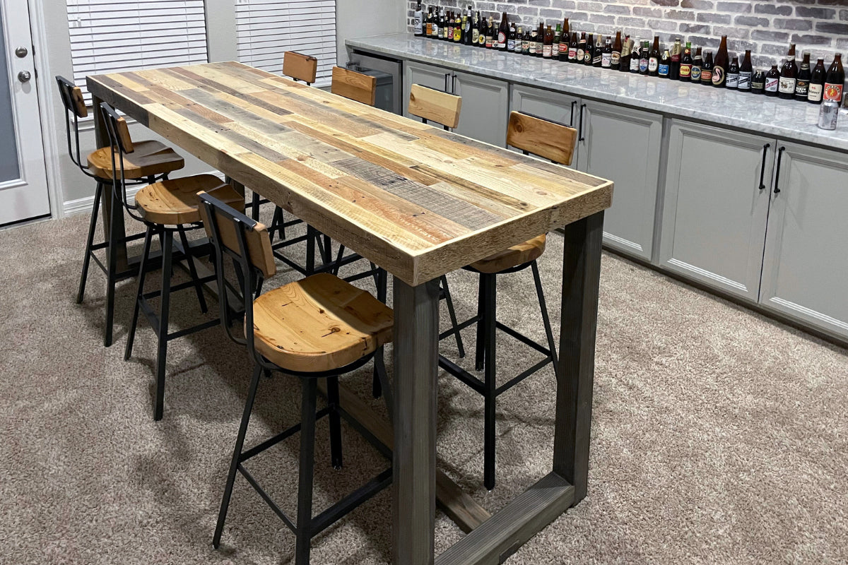 A reclaimed wood bar height table, paired with six wooden and steel chairs in a home bar area. 