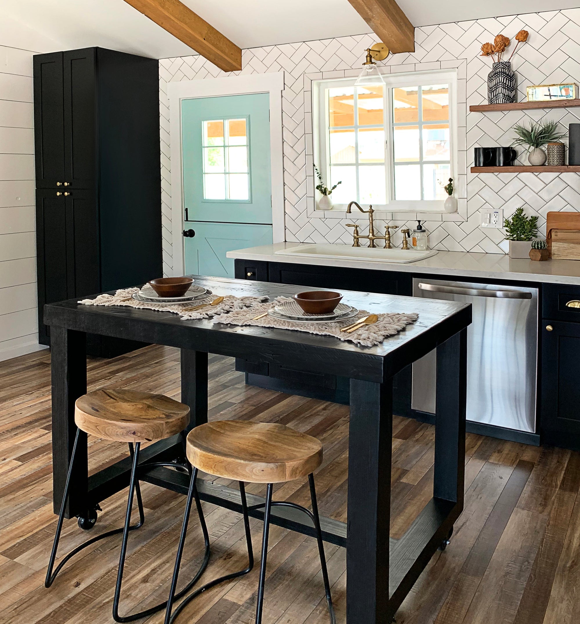 Modern kitchen featuring a handcrafted black kitchen island with two reclaimed wood bar-height stools, set on wood floors with black cabinets, open shelves, and herringbone subway tile backsplash.