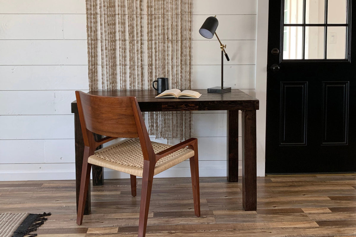 This handcrafted rustic wood desk, built from reclaimed wood and designed for enduring quality, features a chair, open book, black mug, and desk lamp. The scene is set on natural wood flooring beside a black door.