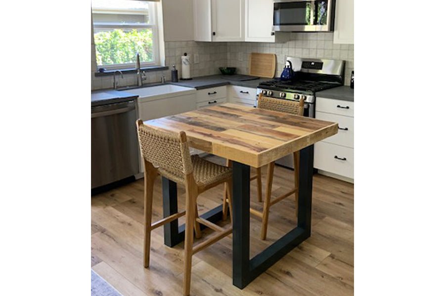 A bright, modern kitchen anchored by a handcrafted Gunnison counter height table featuring industrial steel legs and a reclaimed wood top. Two woven back chairs complete this multi-functional kitchen island setting.