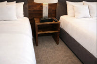 Experience timeless design and exceptional quality with our handcrafted reclaimed wood nightstand, thoughtfully placed between two neatly made hotel beds. This sustainable bedside table features a lamp, clock, and remote for comfort.