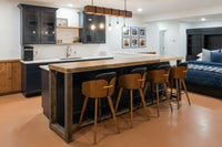 This warm, inviting kitchen showcases a handcrafted reclaimed wood kitchen island with four wooden bar stools. Featuring dark cabinets, open shelves, and pendant lights, this reclaimed wood furniture delivers enduring quality and sustainable style. Framed photos accent the wall while a cozy bed with blue bedding offers a glimpse of adjacent living space.