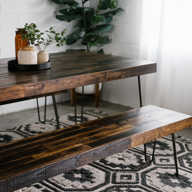 This handcrafted reclaimed wood dining table and matching bench, set atop sleek steel hairpin legs, exemplify enduring quality. Ideal as a narrow restaurant or kitchen table. Accented with plant decor and a soft patterned rug.