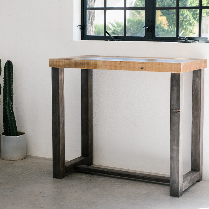 A handcrafted, narrow bar-height console table made with a light reclaimed wood top and dark, sturdy legs stands near a sunlit window. Enduring quality meets sustainable design beside a potted cactus; ideal reclaimed wood furniture.