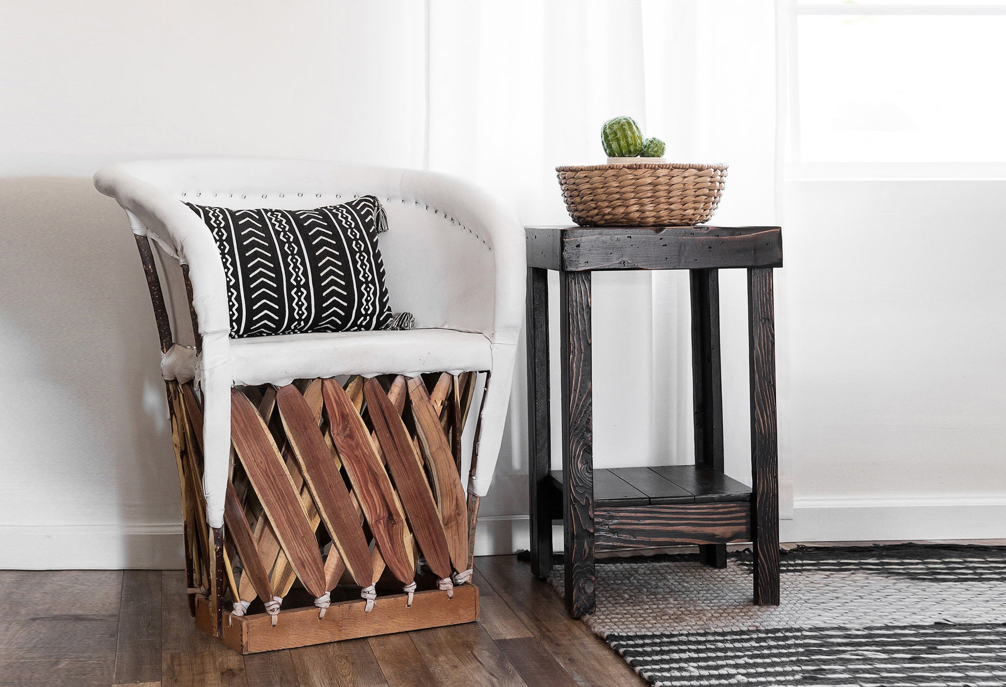 White cushioned chair with black and white patterned pillow beside dark reclaimed wood side table, woven basket with cactus on top; handcrafted furniture on wooden floor by window with white curtains.