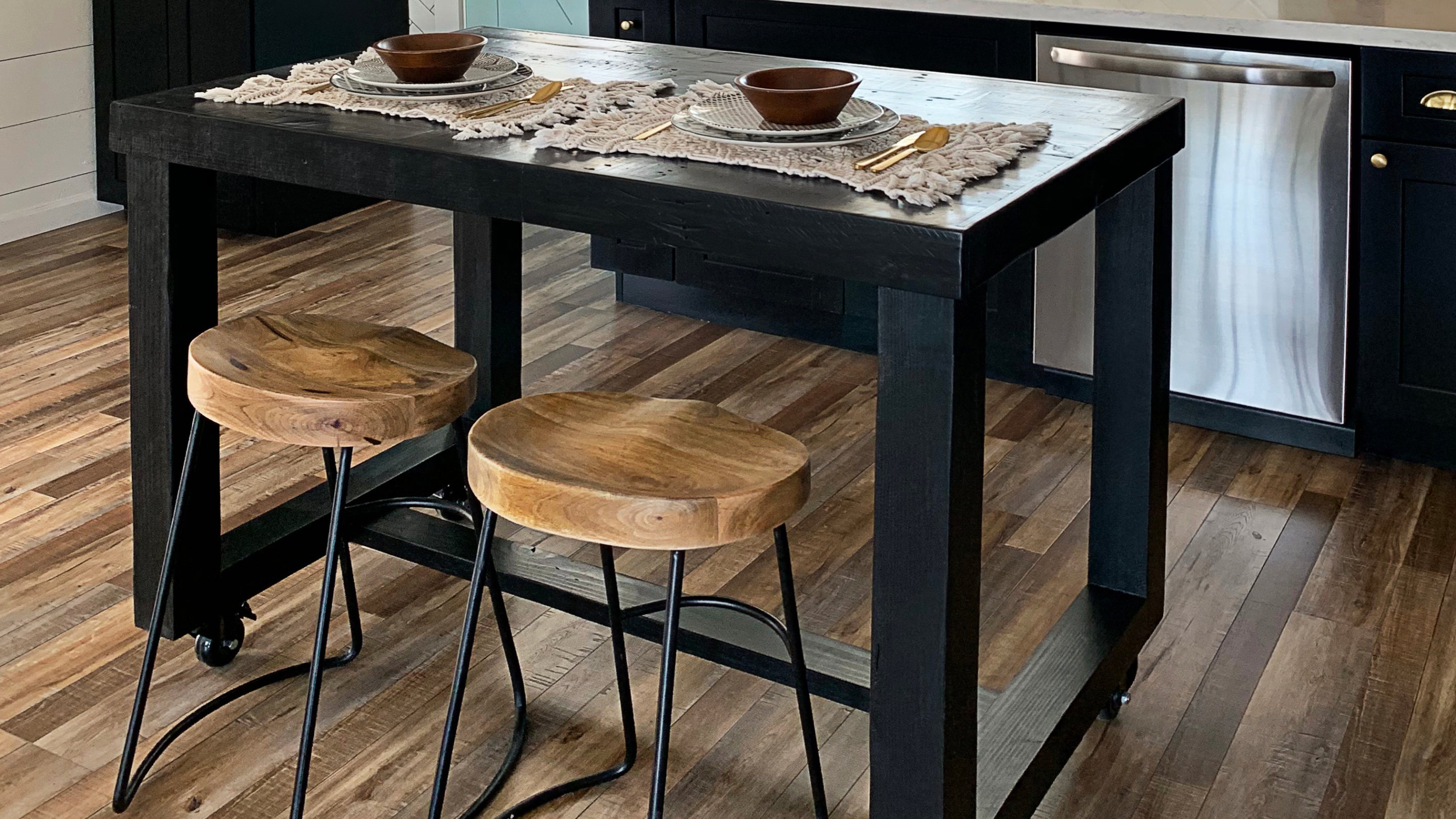 This kitchen showcases a handcrafted black reclaimed wood kitchen island, paired with rustic bar-height stools featuring durable steel legs. Accented with tableware, stainless appliances, and a warm wood floor.
