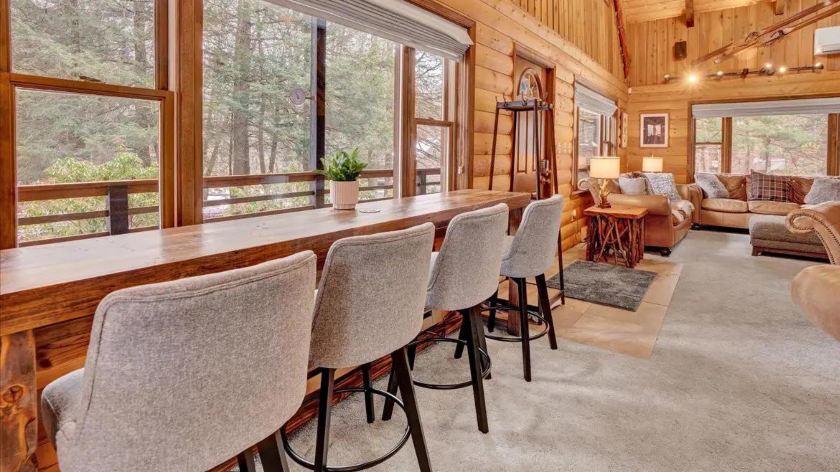 A welcoming log cabin living room features a handcrafted reclaimed wood bar table with four upholstered bar stools, plush sofas, and expansive windows offering serene views of the surrounding trees.