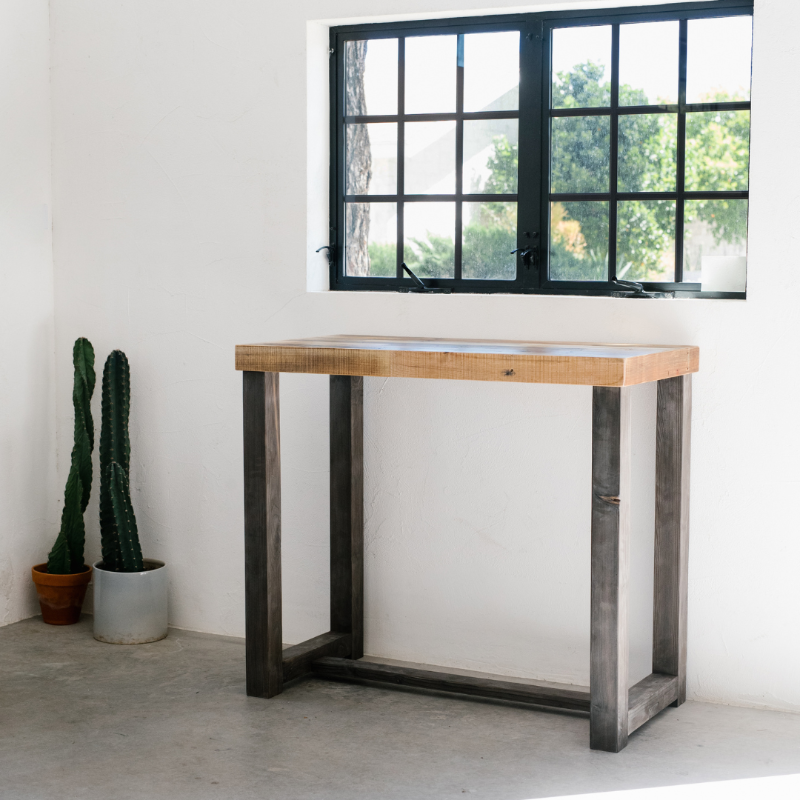 Bar-height dining table crafted from reclaimed wood and steel, set before a black-framed window in a bright minimalist space; two potted cacti accent the furniture’s modern, durable appeal.