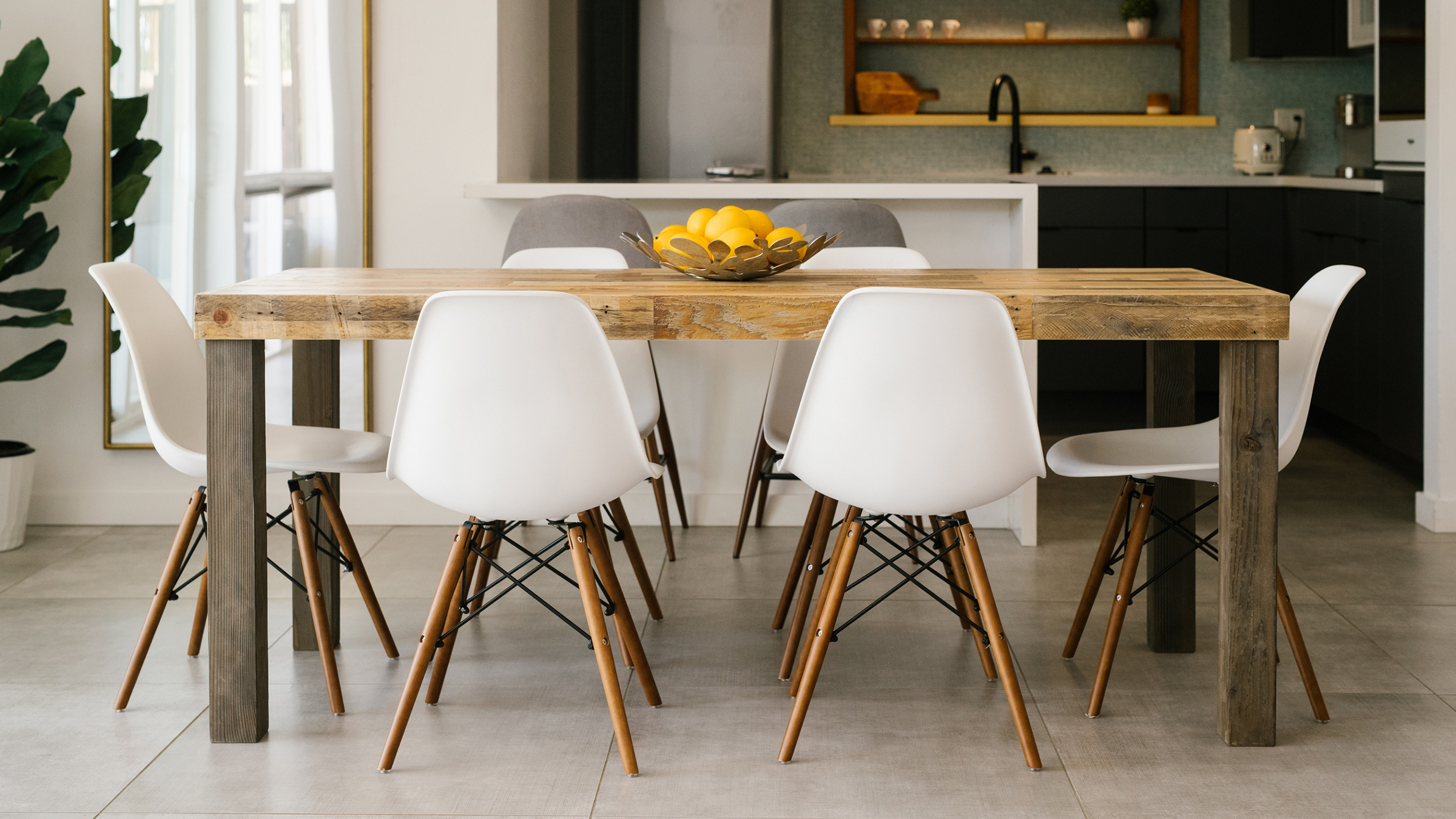 Experience timeless dining in this inviting space featuring a handcrafted reclaimed wood dining table, paired with six white chairs and an artful bowl of lemons. The light floor, indoor plant, and open kitchen complete the welcoming scene.
