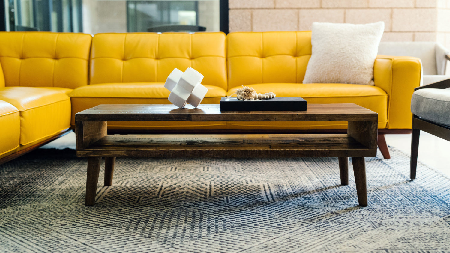 A modern living room showcases a bright yellow sofa with a white pillow, complemented by a handcrafted reclaimed wood coffee table—ideal as a narrow table or restaurant table—accented with decor and set atop a patterned rug.