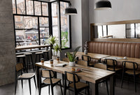 A welcoming cafe with large windows features a handcrafted bar-height table made from reclaimed wood, complemented by black chairs and brown cushioned benches. Warm pendant lighting highlights enduring craftsmanship and sustainable materials.