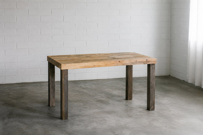 Solid reclaimed wood desk with grey legs against a white brick wall. Handmade by Kase Custom.