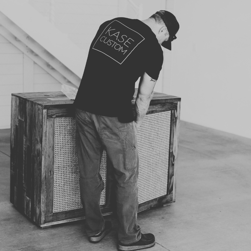 Black and white photo of a man wearing a shirt with the Kase Custom logo on it, tending to hardware of a piece of custom wood furniture