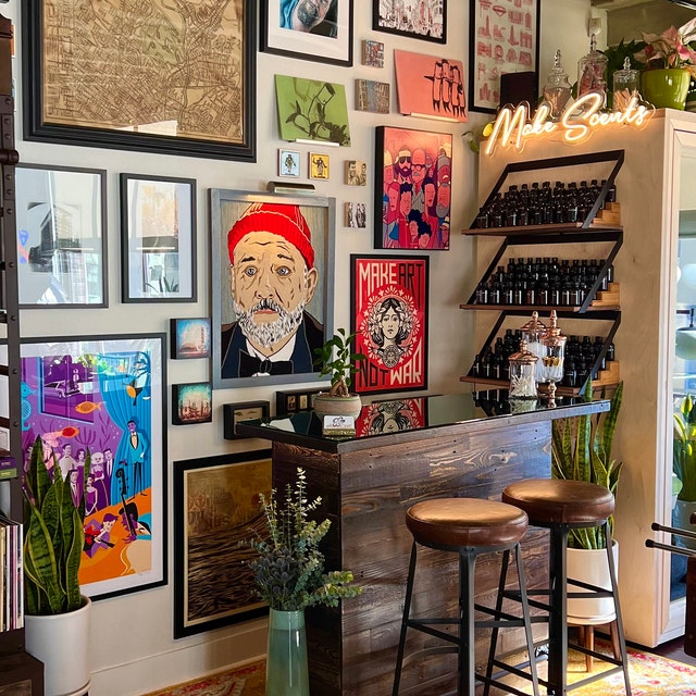 Cozy shop corner featuring a handcrafted reclaimed wood counter with two bar-height stools, potted plants, shelves of bottles, and colorful art on the walls beneath a Make Scents neon sign.
