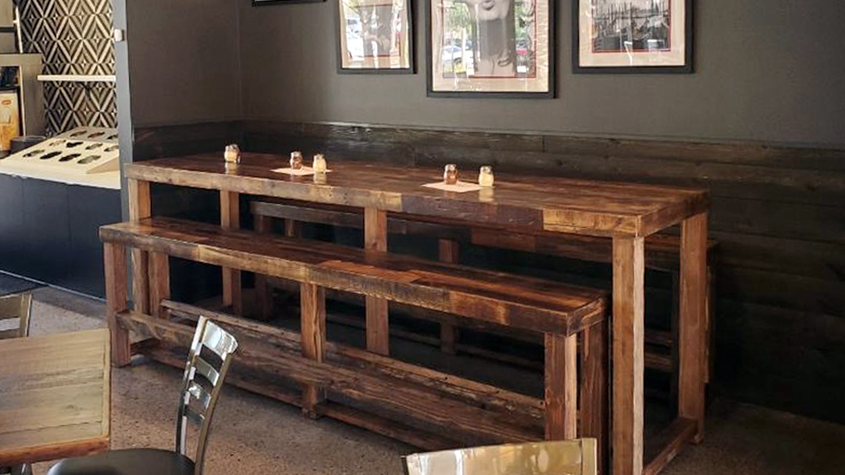 A handcrafted reclaimed wood restaurant bar table with matching benches stands against a dark cafe wall, embodying timeless craftsmanship. Three black-and-white photos and candles accent this sustainable restaurant table design.
