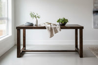 This handcrafted reclaimed wood bar-height table in Espresso is perfect as a kitchen island or dining table, features enduring quality and sustainable materials. Styled with a white vase, plates, and a green plant in a minimalistic room.