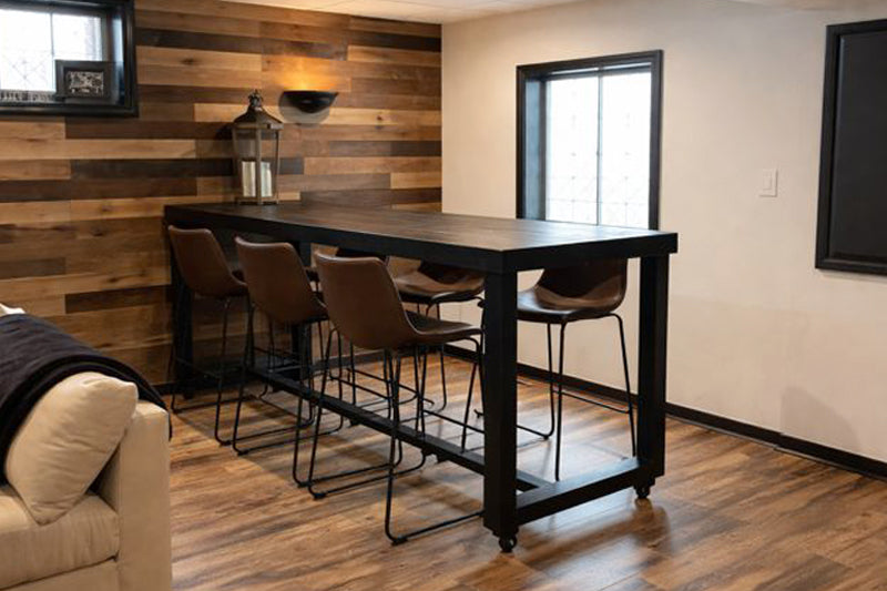 This inviting dining area showcases a handcrafted bar-height table made from reclaimed wood, paired with five brown leather bar stools. The warm wood-paneled accent wall and hardwood floors highlight enduring quality and sustainable design.