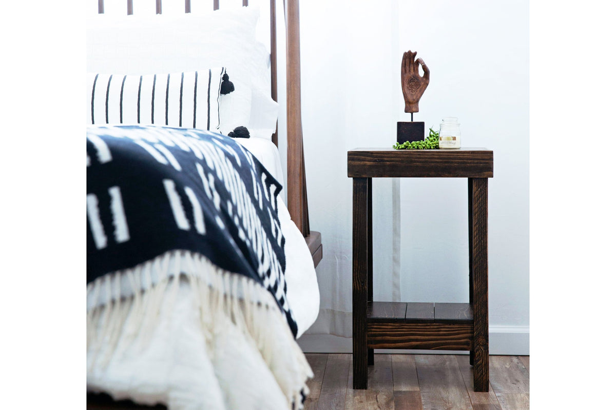 Handcrafted in Arizona, this reclaimed wood nightstand showcases enduring quality beside a bed dressed in a black-and-white patterned blanket. A hand-shaped sculpture, plant, and candle add warmth to the serene setting—perfect as sustainable bedroom furniture.