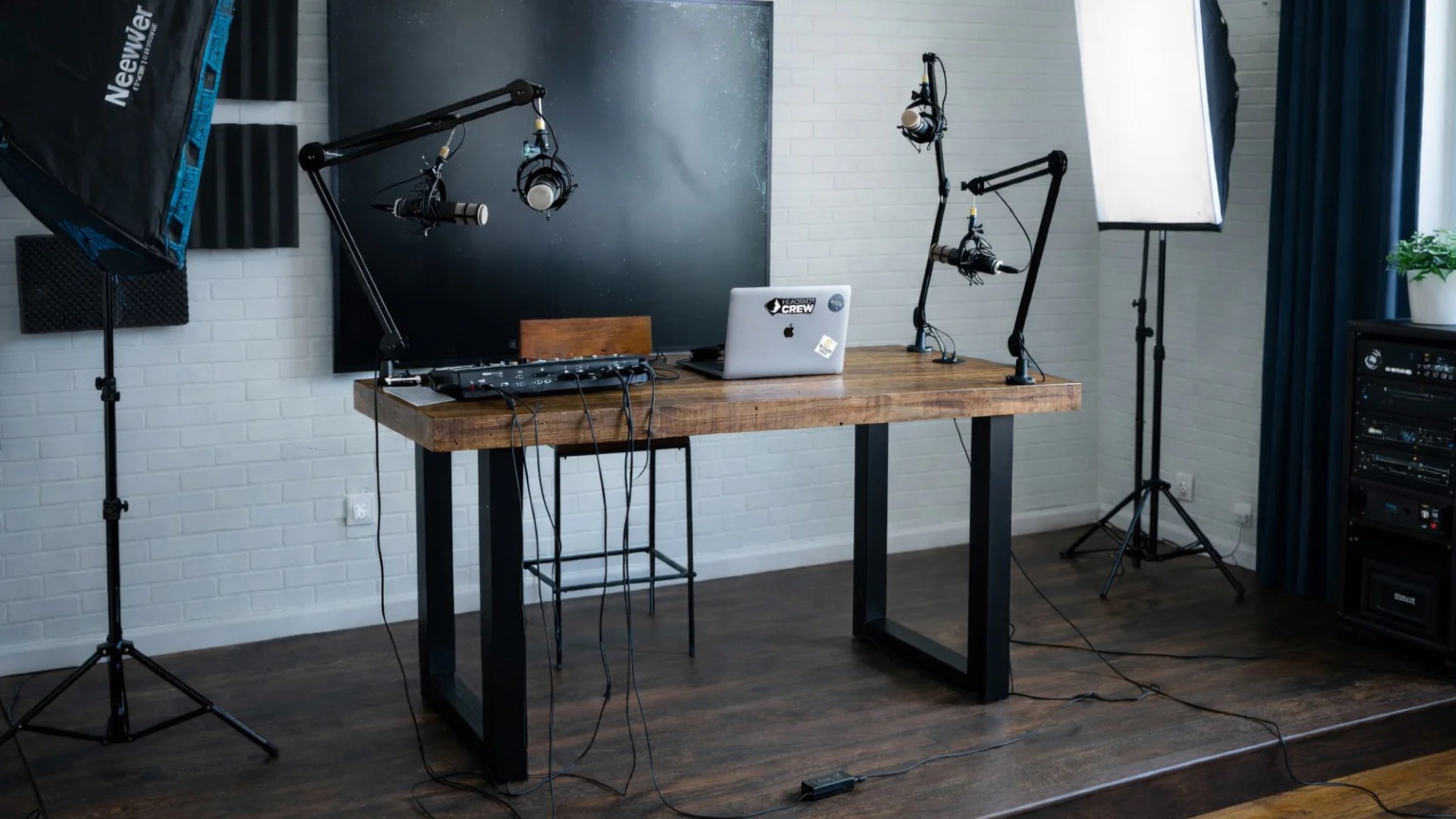 A bright, professional podcast studio features a handcrafted reclaimed wood bar table with steel legs, microphones on adjustable arms, headphones, a laptop, audio gear, blackboard, and photography lighting set against white brick walls.