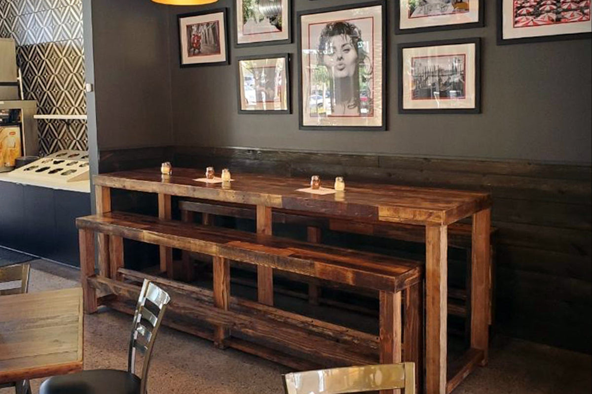 A welcoming café corner features handcrafted reclaimed wood bar tables and a reclaimed wood bench, perfect as restaurant tables or bar-height seating. Warm lighting and artisanal décor highlight enduring quality and sustainable design.