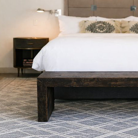 A modern bedroom featuring a white bed with patterned pillows, complemented by a handcrafted dark wood bench at the foot. A geometric rug anchors the space, paired with a reclaimed wood nightstand for timeless quality.