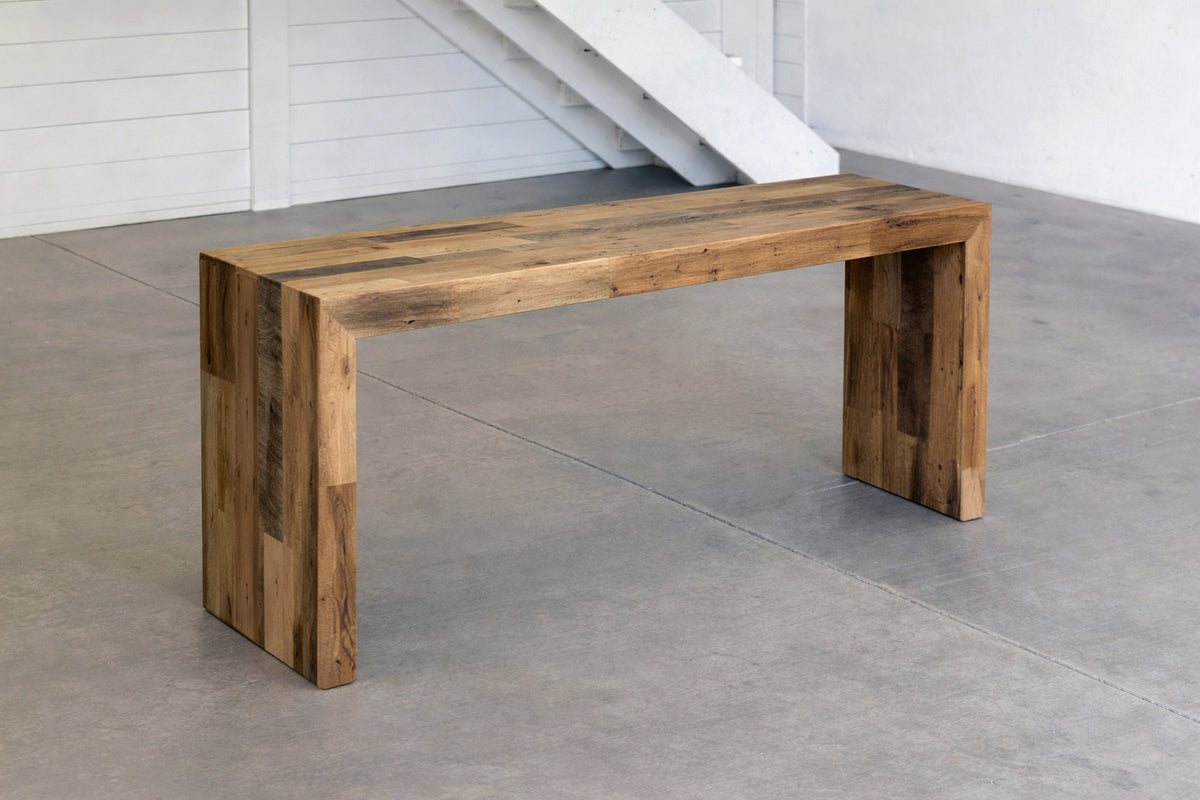 Handcrafted reclaimed wood console table with modern waterfall sides in a minimalistic indoor setting with concrete floor and white walls.