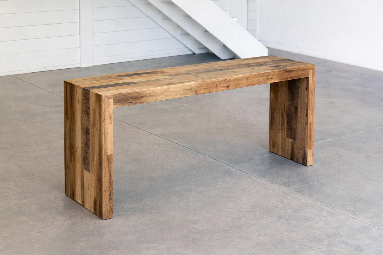 Handcrafted reclaimed wood bench with modern waterfall sides in a minimalistic indoor setting with concrete floor and white walls.