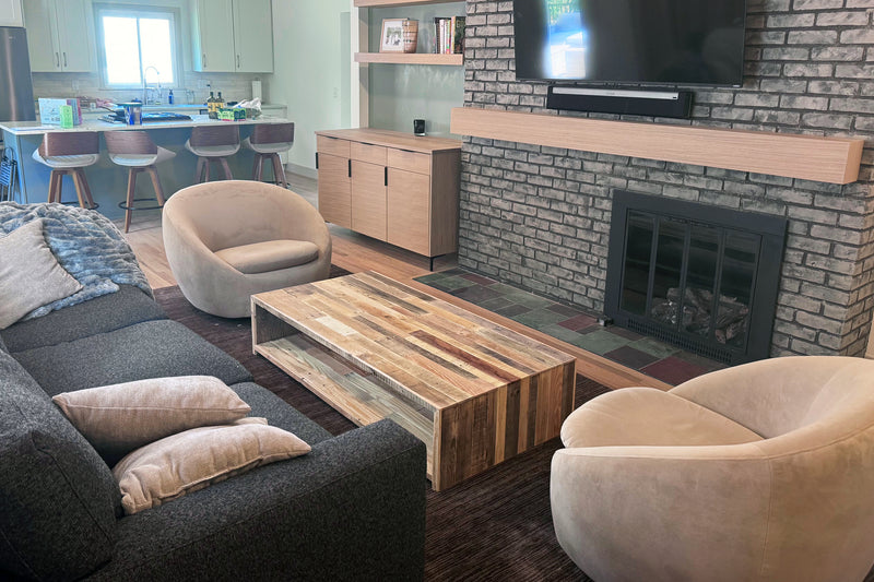 Modern living room featuring a dark sofa, two beige armchairs, and a handcrafted rectangular coffee table made from reclaimed wood. Fireplace with TV above and kitchen with bar stools in the background.
