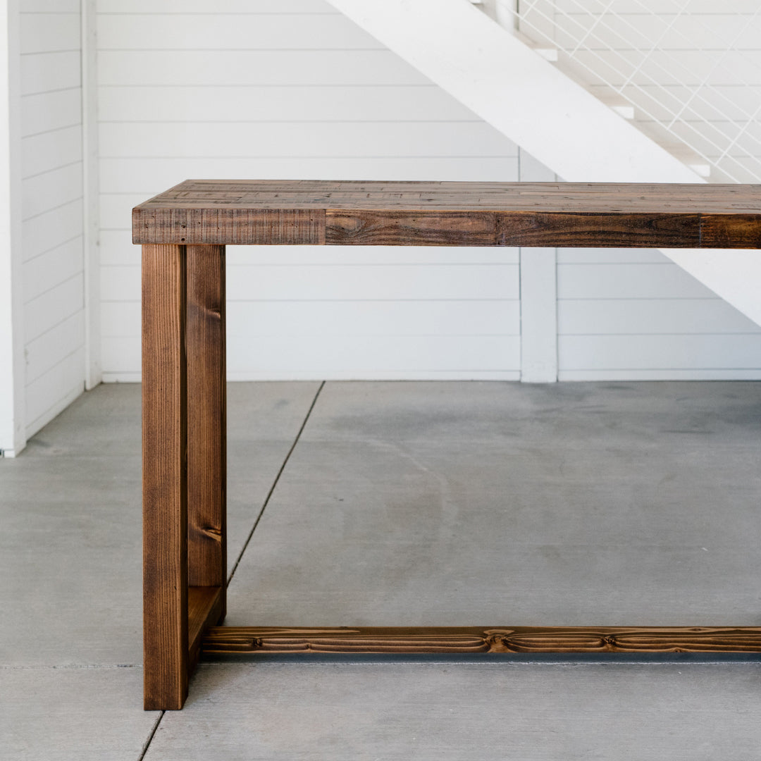Experience the timeless appeal of our handcrafted bar-height table, expertly made in Arizona from reclaimed wood with a rich dark brown finish. Ideal as a narrow kitchen island or restaurant table, it highlights enduring quality and sustainable materials against crisp white paneled walls and a modern staircase backdrop.