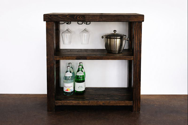 Expertly crafted from dark reclaimed wood, this sustainable bar cart features two shelves—perfect as a narrow bar table or kitchen island. The top shelf holds wine glasses and an ice bucket; the lower displays sparkling water.