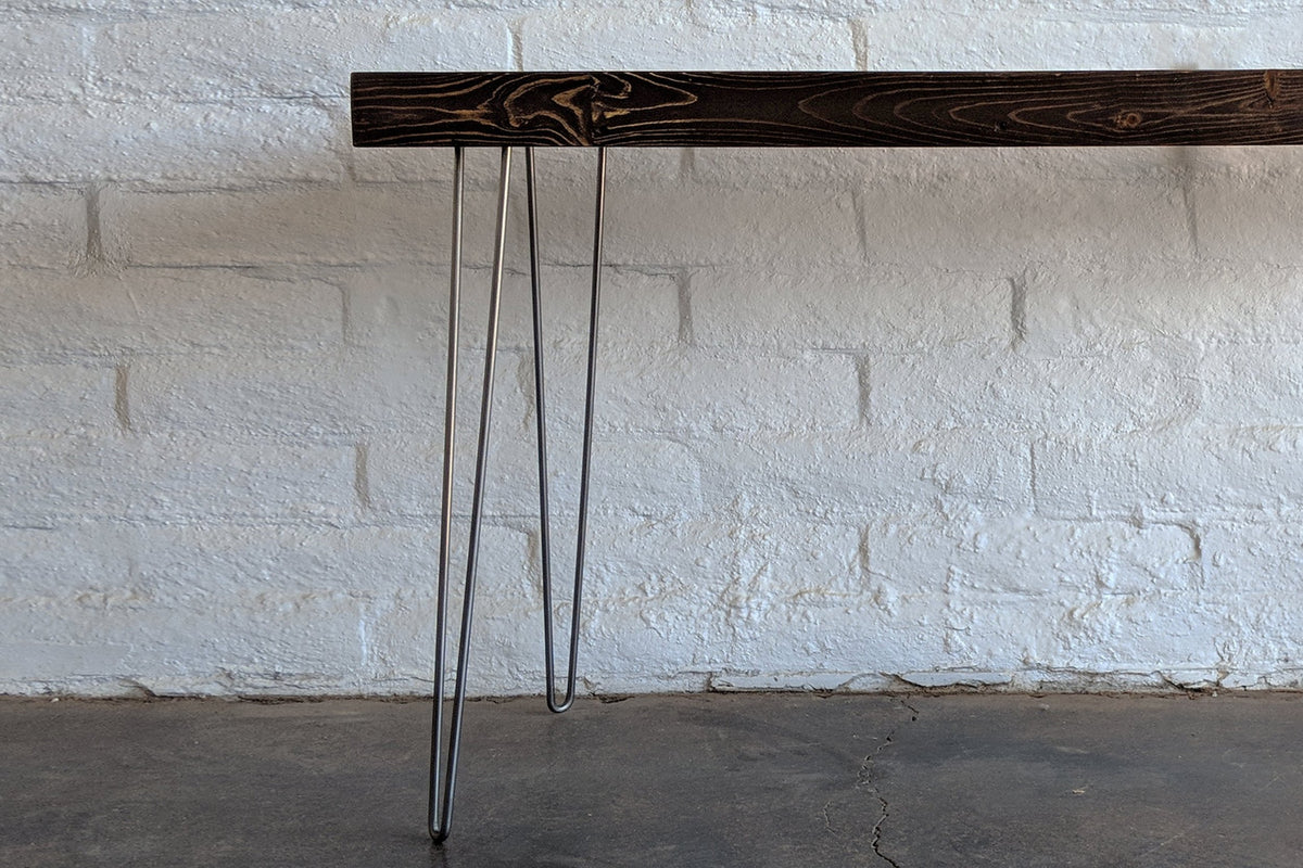 A close-up of a handcrafted reclaimed wood desk with sleek steel hairpin legs, set against a white brick wall and concrete floor. Sustainable materials highlight enduring quality.