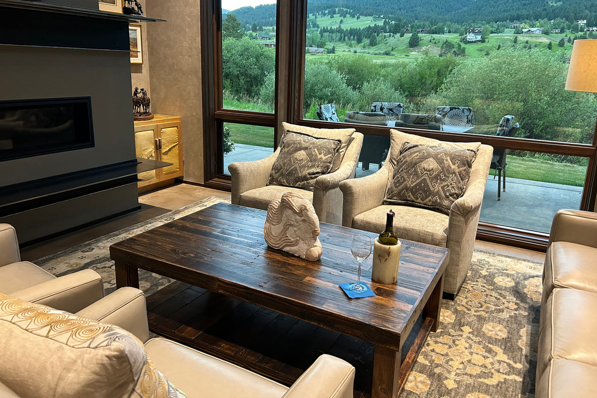 A warm, inviting living room features beige armchairs and sofas around a handcrafted reclaimed wood coffee table. Large windows frame stunning mountain and greenery views.
