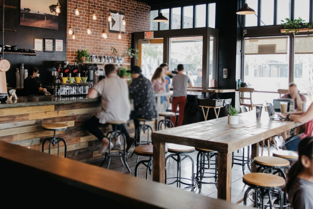 Bar-height dining table and communal table handcrafted in Arizona from reclaimed wood and steel, featured in a cozy coffee shop with exposed brick walls, hanging lights, and large windows. Ideal for modern café or historic home settings.