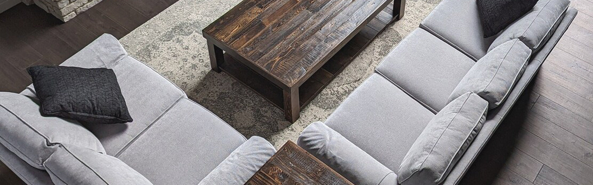Top-down view of a living room featuring two handcrafted light gray sofas with black cushions, a reclaimed wood coffee table, and a textured area rug on dark wood flooring—ideal for modern or historic homes.