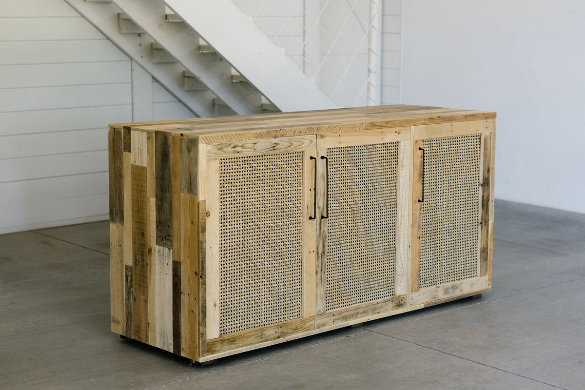 Handcrafted wooden sideboard with three cane panel doors and metal handles, shown on a concrete floor beside a white staircase. Ideal for modern minimalist spaces. Sustainable materials. Arizona-made furniture.