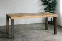 A rustic rectangular wooden dining table made from reclaimed wood, set against a white brick wall and concrete floor.