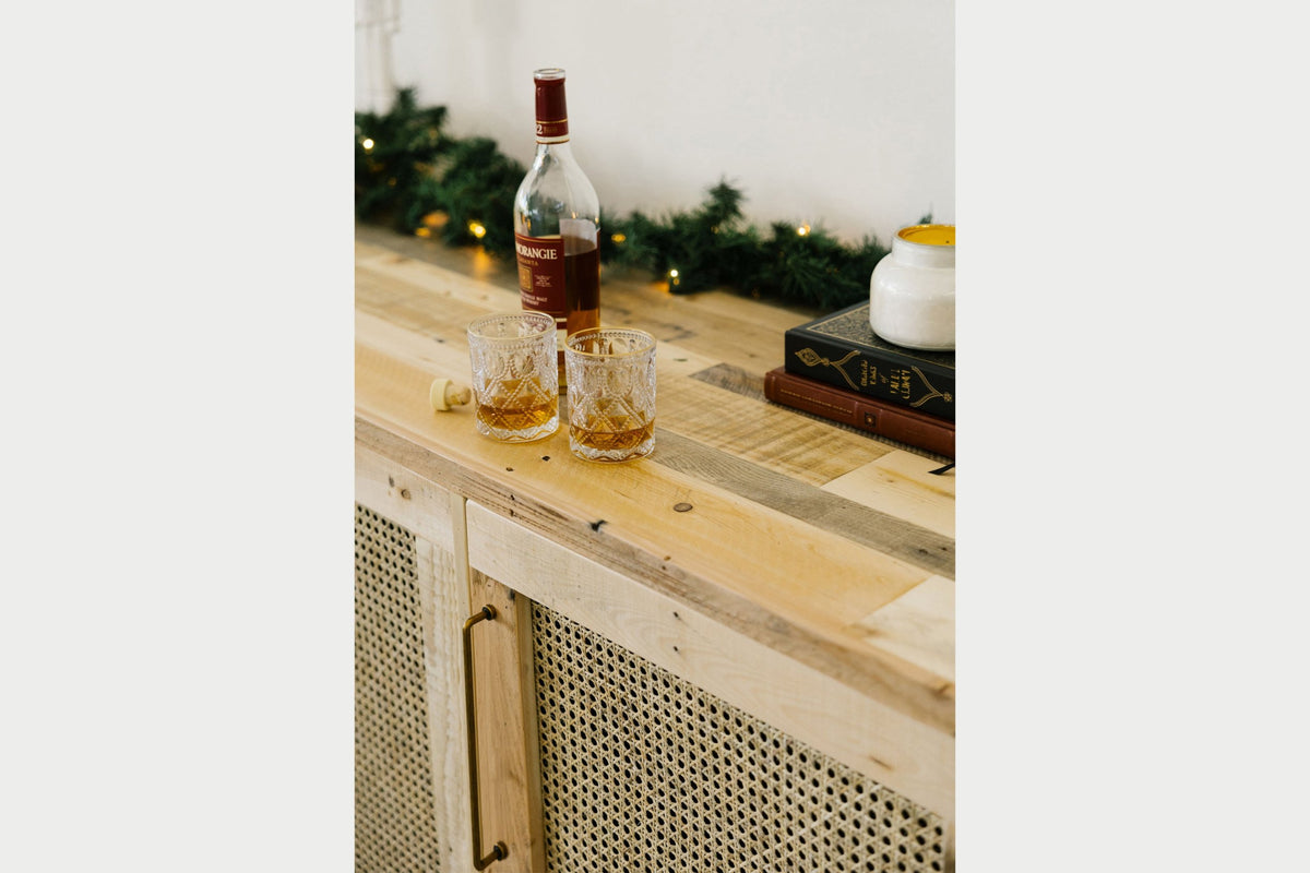 A bottle of whiskey, two filled glasses, a cork, a candle, and stacked books are elegantly arranged on a handcrafted reclaimed wood sideboard.