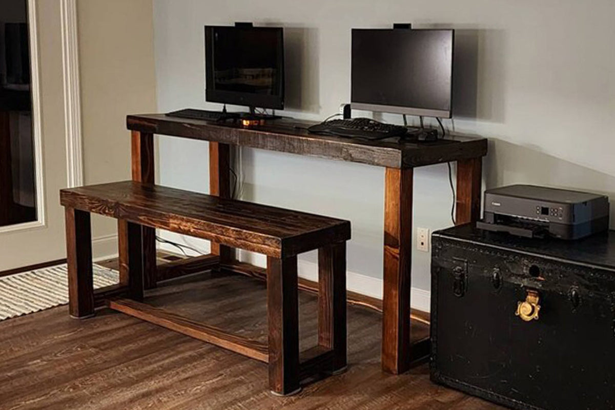 A handcrafted reclaimed wood desk serves as a spacious workspace with dual monitors, keyboard, and mouse, complemented by a counter-height reclaimed wood bench. Ideal as a narrow bar-height table or kitchen island; black trunk and printer nearby.