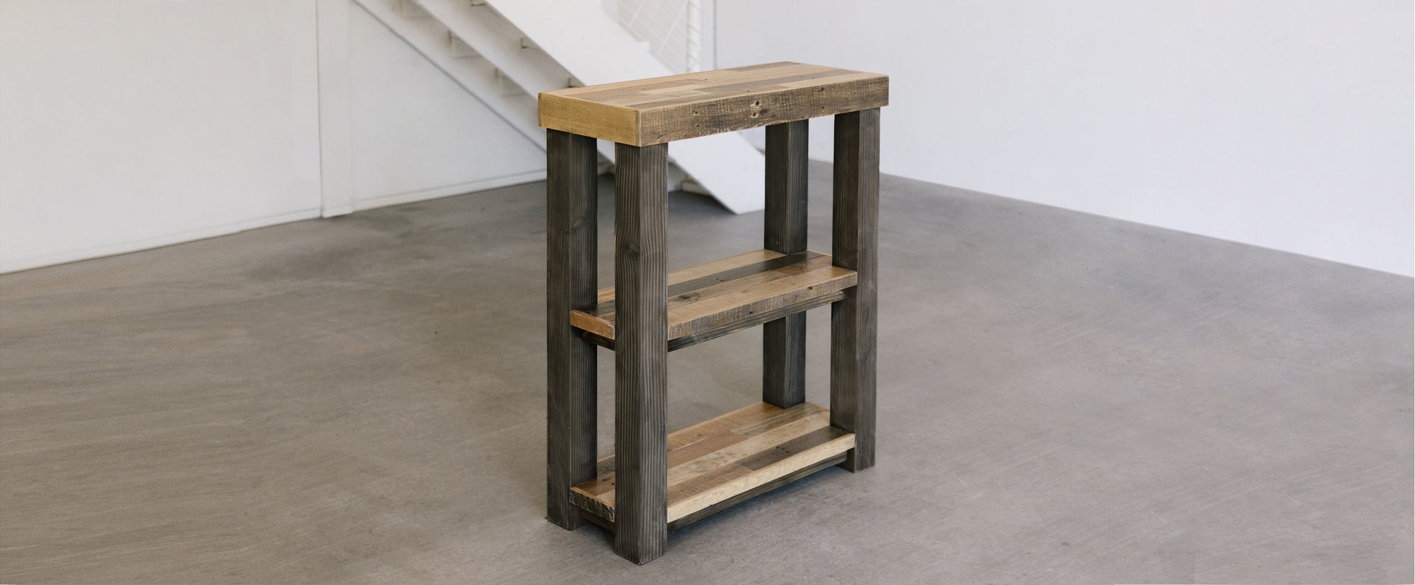 Rustic side table, handcrafted in Arizona from reclaimed wood planks, features two open shelves. Ideal for minimalist spaces or historic homes. Shown on a smooth gray floor by a white wall and staircase.