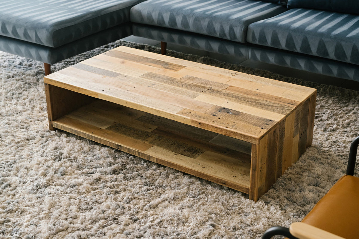 This handcrafted reclaimed wood coffee table features an open shelf, showcasing enduring quality and artisan craftsmanship. Perfect for modern living, it sits atop a textured beige carpet before a dark gray sectional sofa.