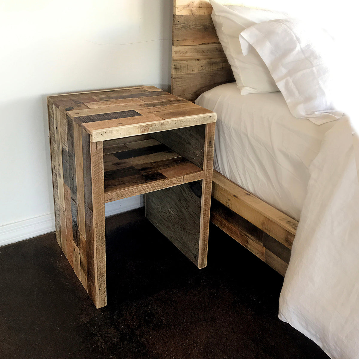 Handcrafted reclaimed wood nightstand with patchwork design and open shelf beside bed with white bedding; matching wooden bed frame on dark floor and white wall.