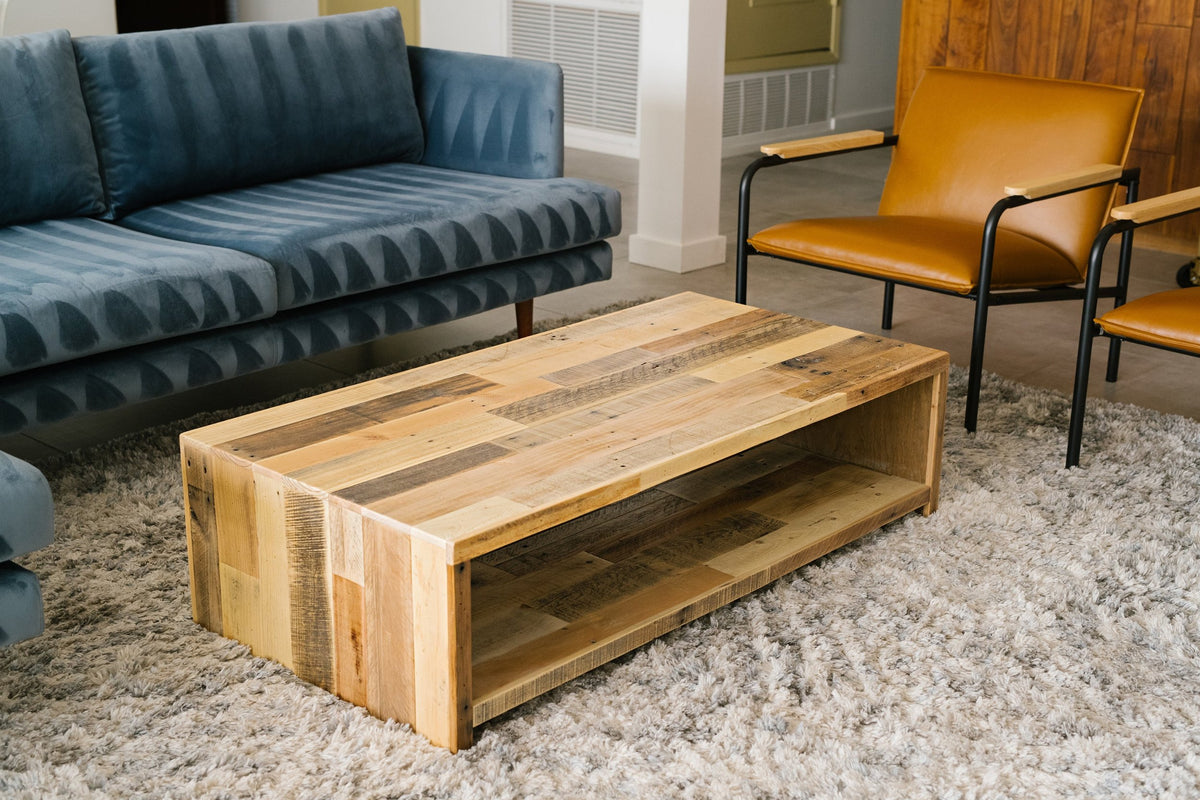 Modern living room featuring a blue sofa, two brown leather chairs with steel arms, and a handcrafted reclaimed wood coffee table in natural finish on a textured gray rug. Sustainable furniture for contemporary spaces.