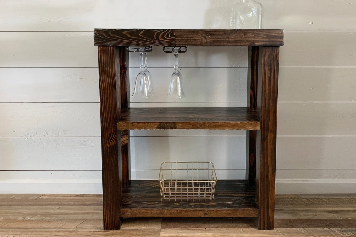 Handcrafted in Arizona, this rustic bar cart showcases reclaimed wood and a sturdy steel frame. Two hanging wine glasses and a metal wire basket add timeless style.